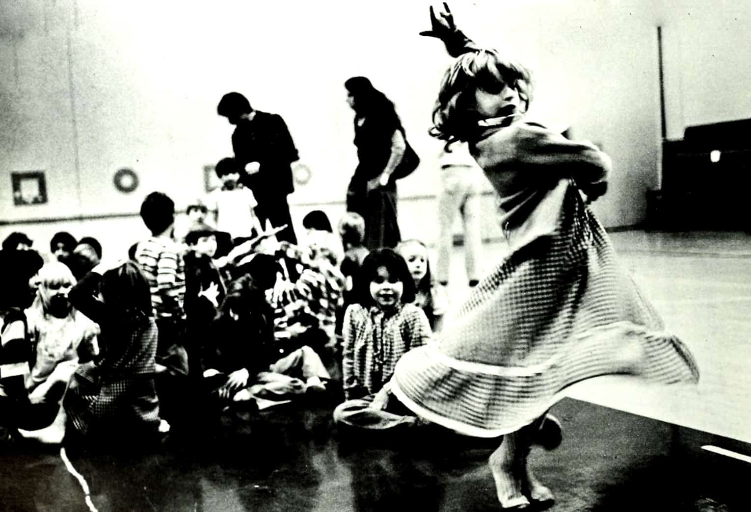 Kids and a dancer in a community center or gymnasium, engaging in a dance or performance, with a lively, energetic atmosphere; black and white photo capturing childhood joy and cultural activities.