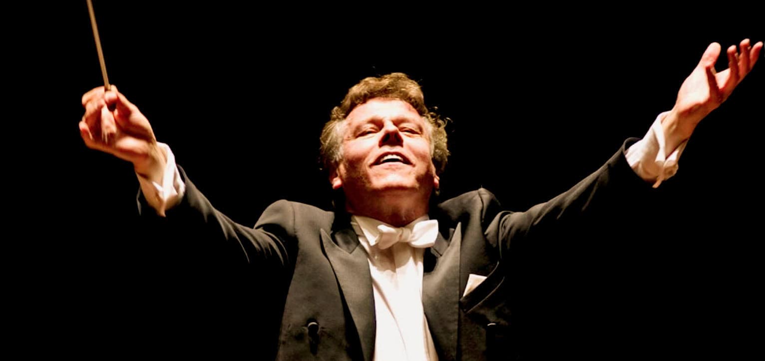 Classic male conductor in tuxedo passionately leading an orchestra with arms raised during a concert, emphasizing musical passion and live performance, with cinematic lighting.