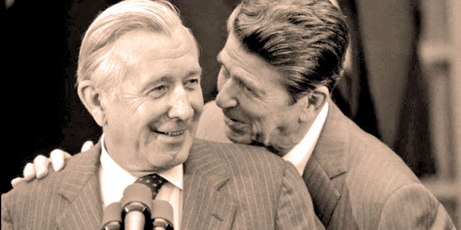 Historic black and white photo of two men in suits, one whispering to the other during a press conference or event. Focus on news, history, and music archive content from Past Daily.