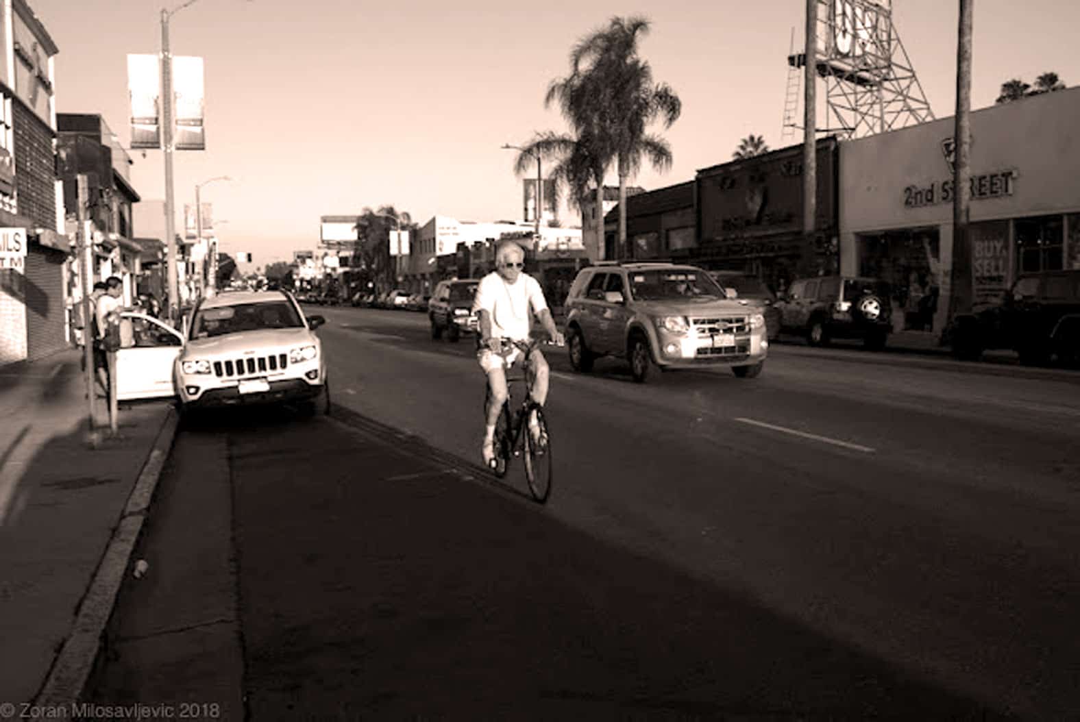 Bike rider on urban street with parked cars and retail shops, vintage sepia tone, capturing city life, music, news and history moments, as featured on Past Daily: A Sound Archive of News, History And Music.