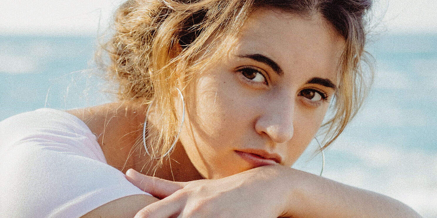 Close-up of a young woman with wavy blonde hair resting her head on her arm at the beach, highlighting a relaxed and summery vibe, perfect for beach lifestyle and serene scenery themes.