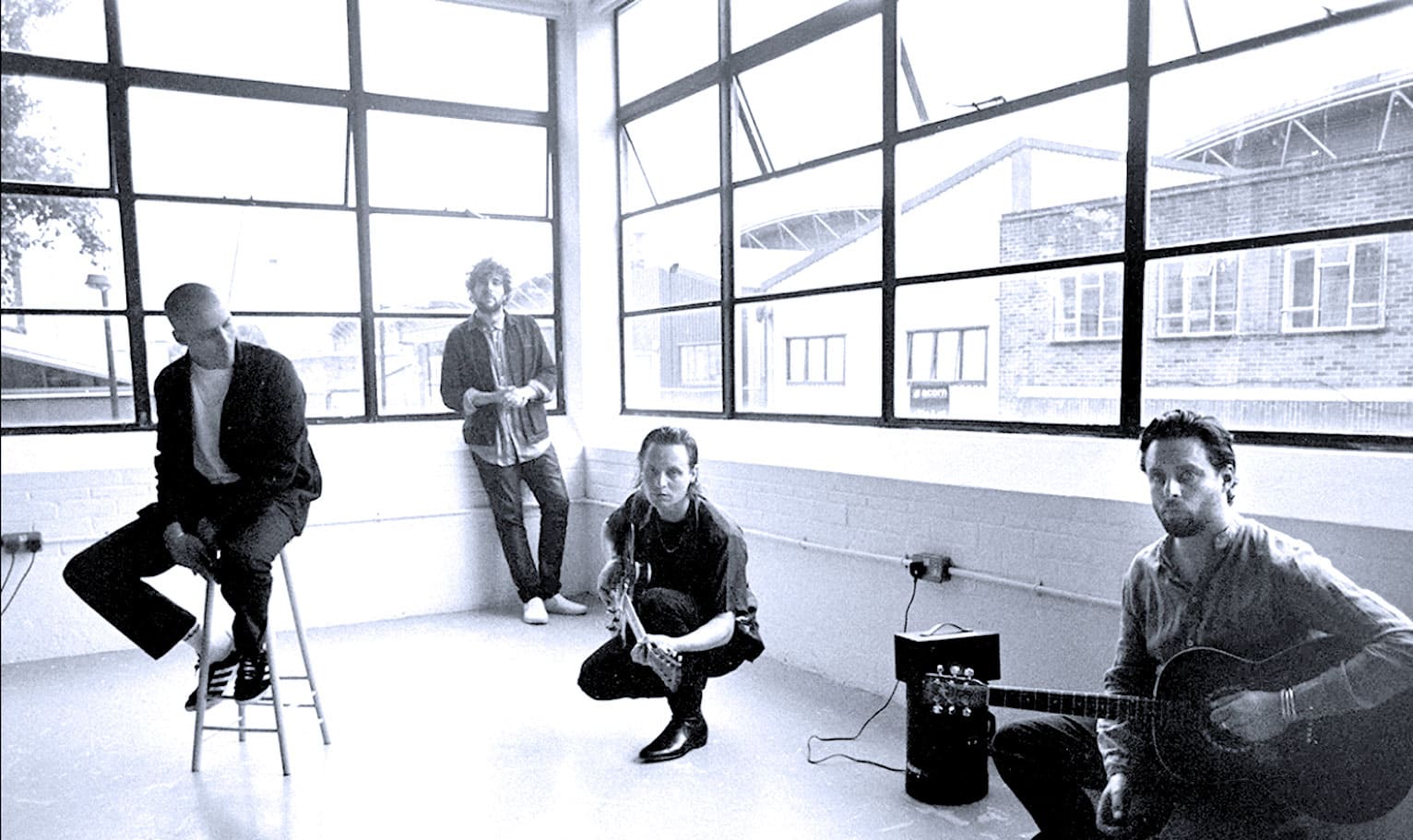 A black and white photo of four musicians in a modern studio with large windows, featuring a guitarist, a woman with a tambourine, and two men, one sitting on a stool. The image emphasizes music, recording, and sound archive themes.