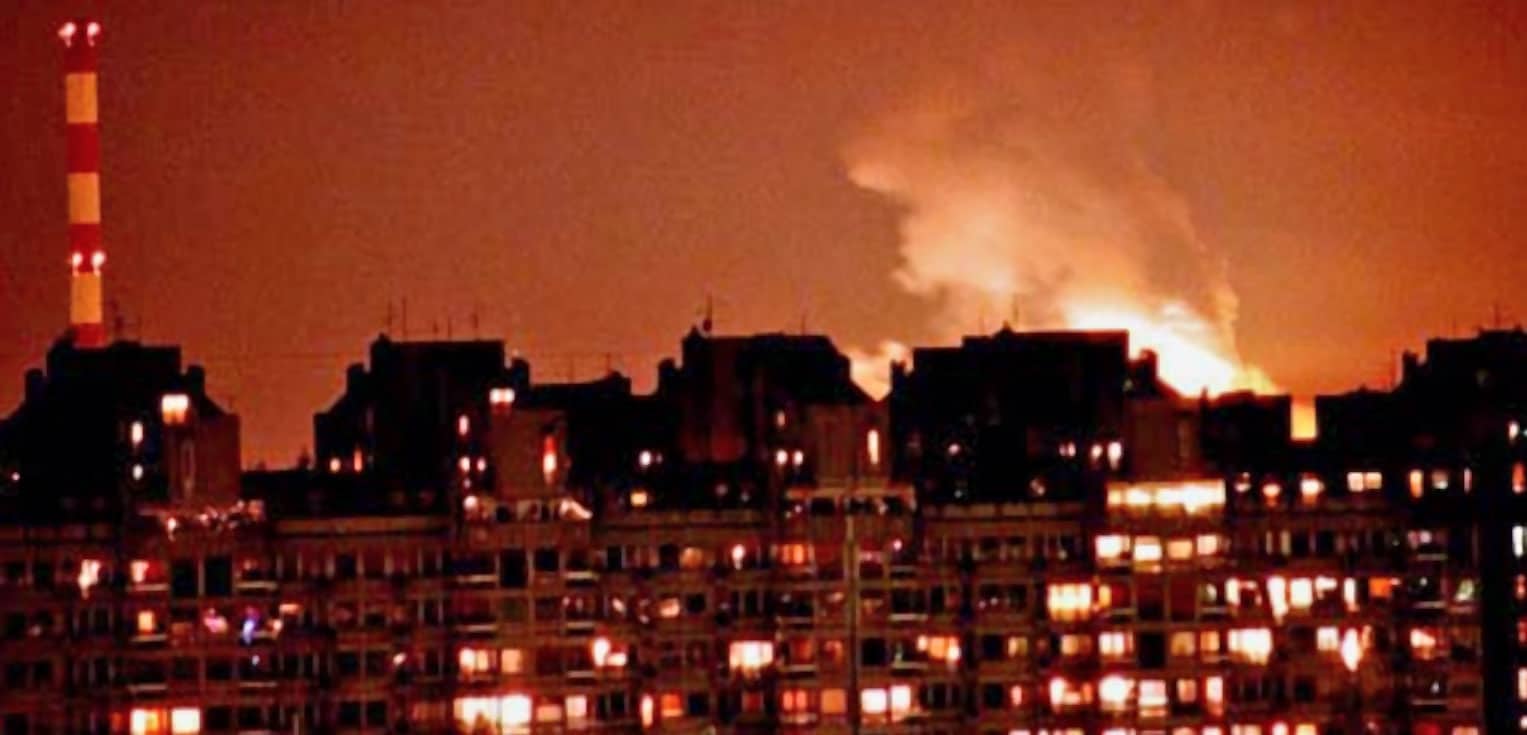 Flickering lights in a city skyline at night with smoke rising from buildings, depicting urban environment, related to news, history, and audio archives of past events.