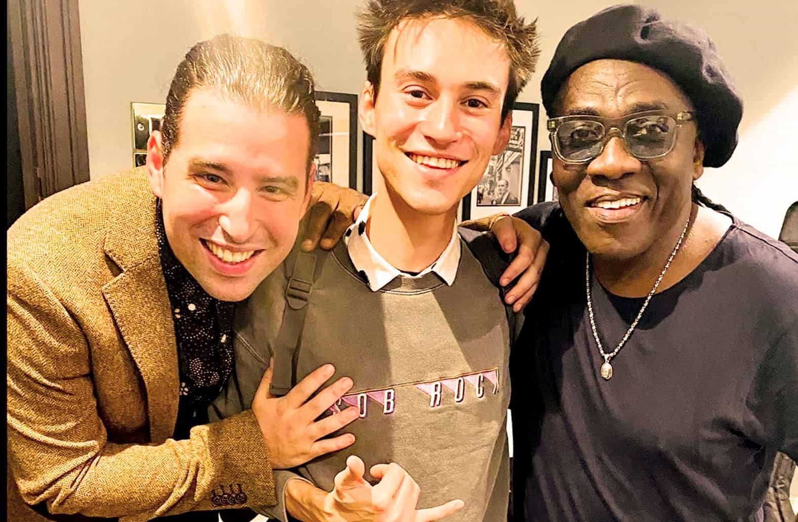 Lively group of three men smiling and posing together at a music or news event, reflecting a sense of camaraderie and celebration, with a background showcasing framed photos, emphasizing music and media culture.
