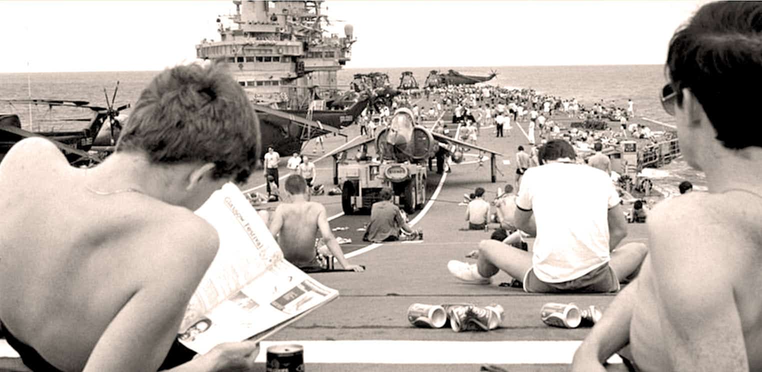 A sound archive of historical news, music, and events featuring a vintage navy aircraft carrier during a seaside gathering with people relaxing and enjoying the maritime setting.