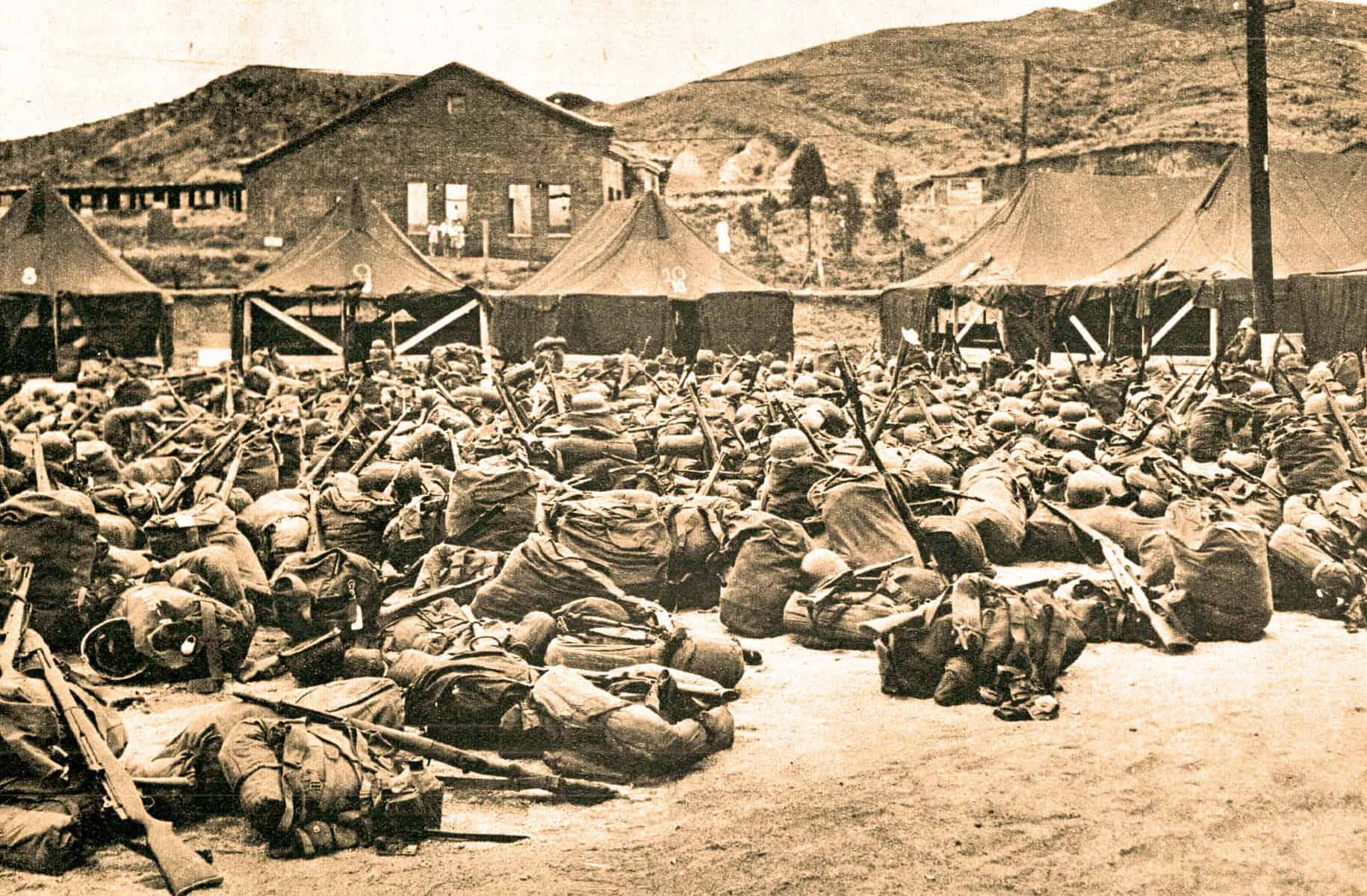 Soldiers kneeling and lying on the ground during military training or drill exercises in front of tents. Historic black-and-white photograph with barracks and hilly landscape in the background, reflecting wartime or military history themes.