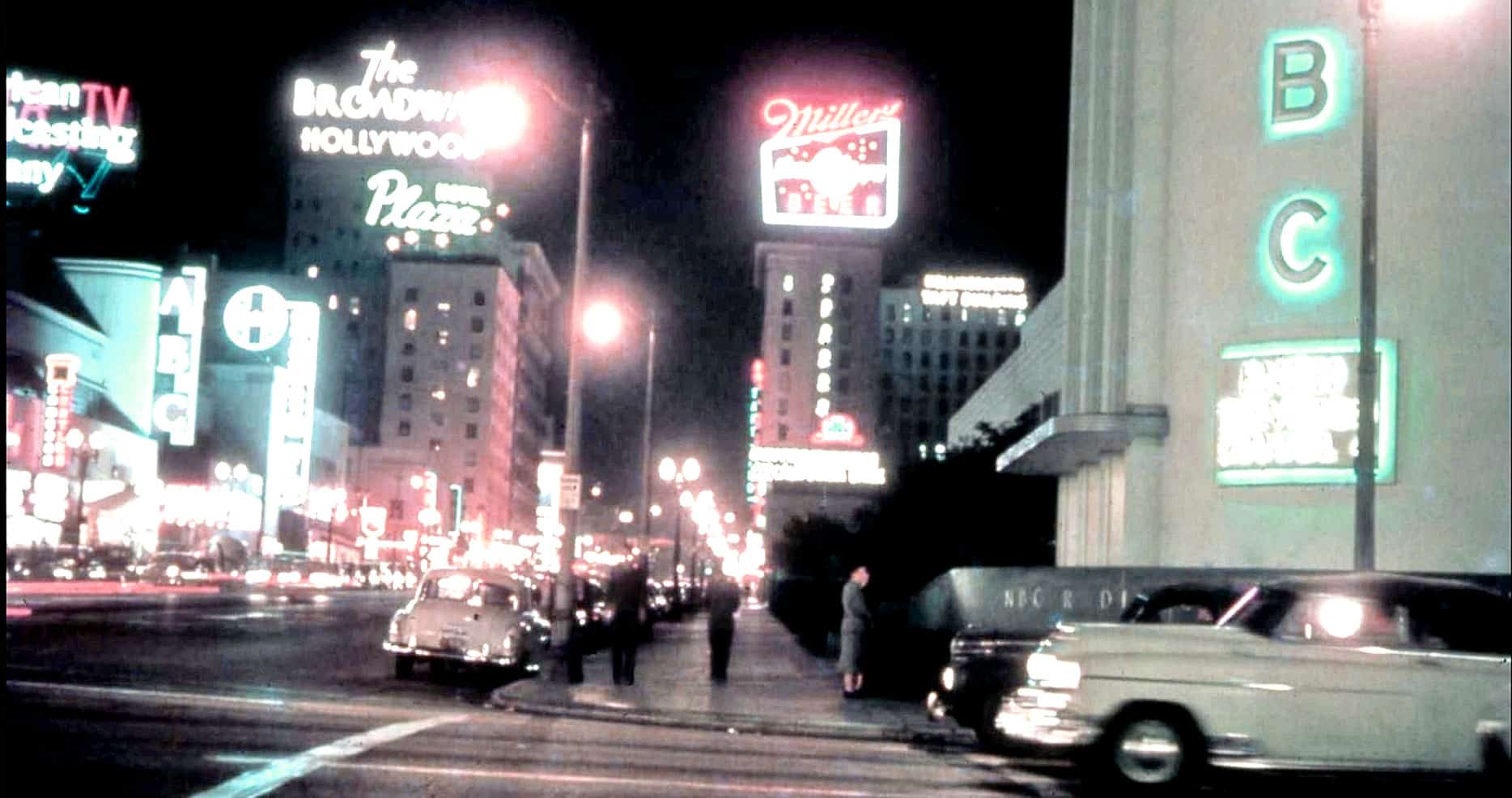 Night city street with neon signs, vintage cars, and pedestrians, showcasing classic Hollywood nightlife, music, entertainment, and urban atmosphere from the mid-20th century.