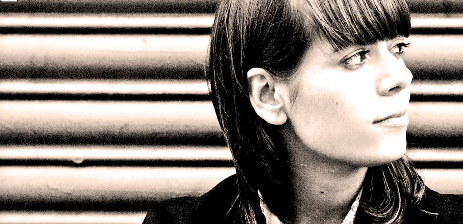 A woman with dark hair and bangs looking thoughtfully to the right with a striped background, representing historical news and music archives.