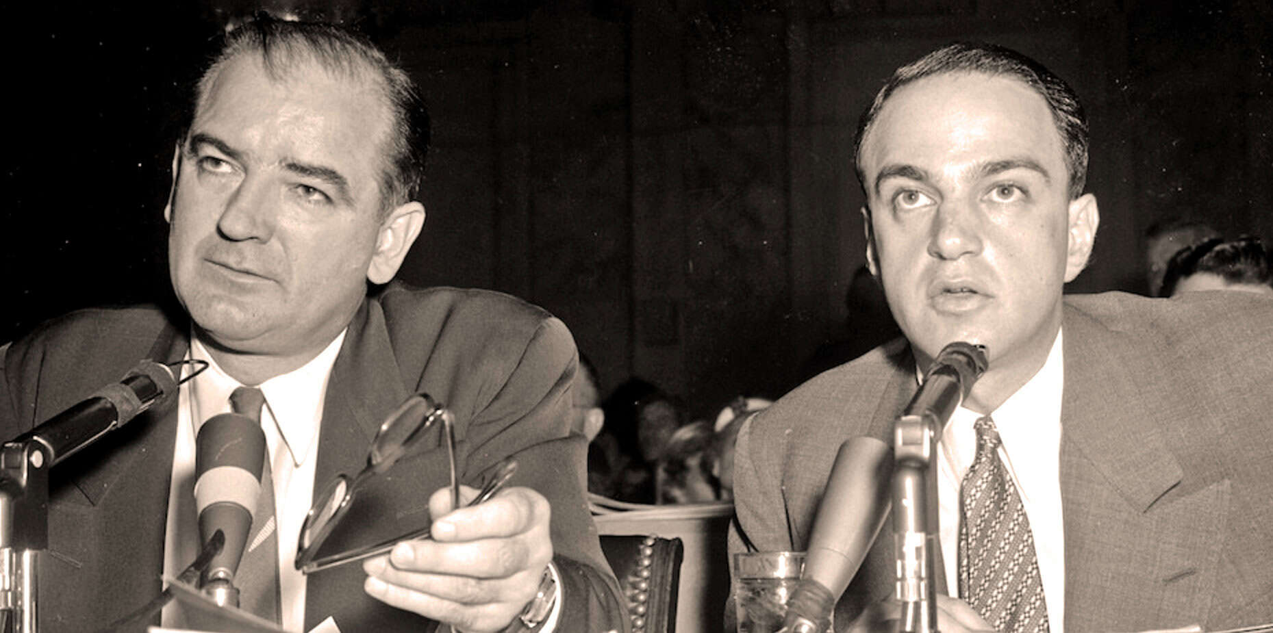 Vintage black and white photo of two men at a 1950s press conference, with microphones in front of them, representing news, history, and media coverage.