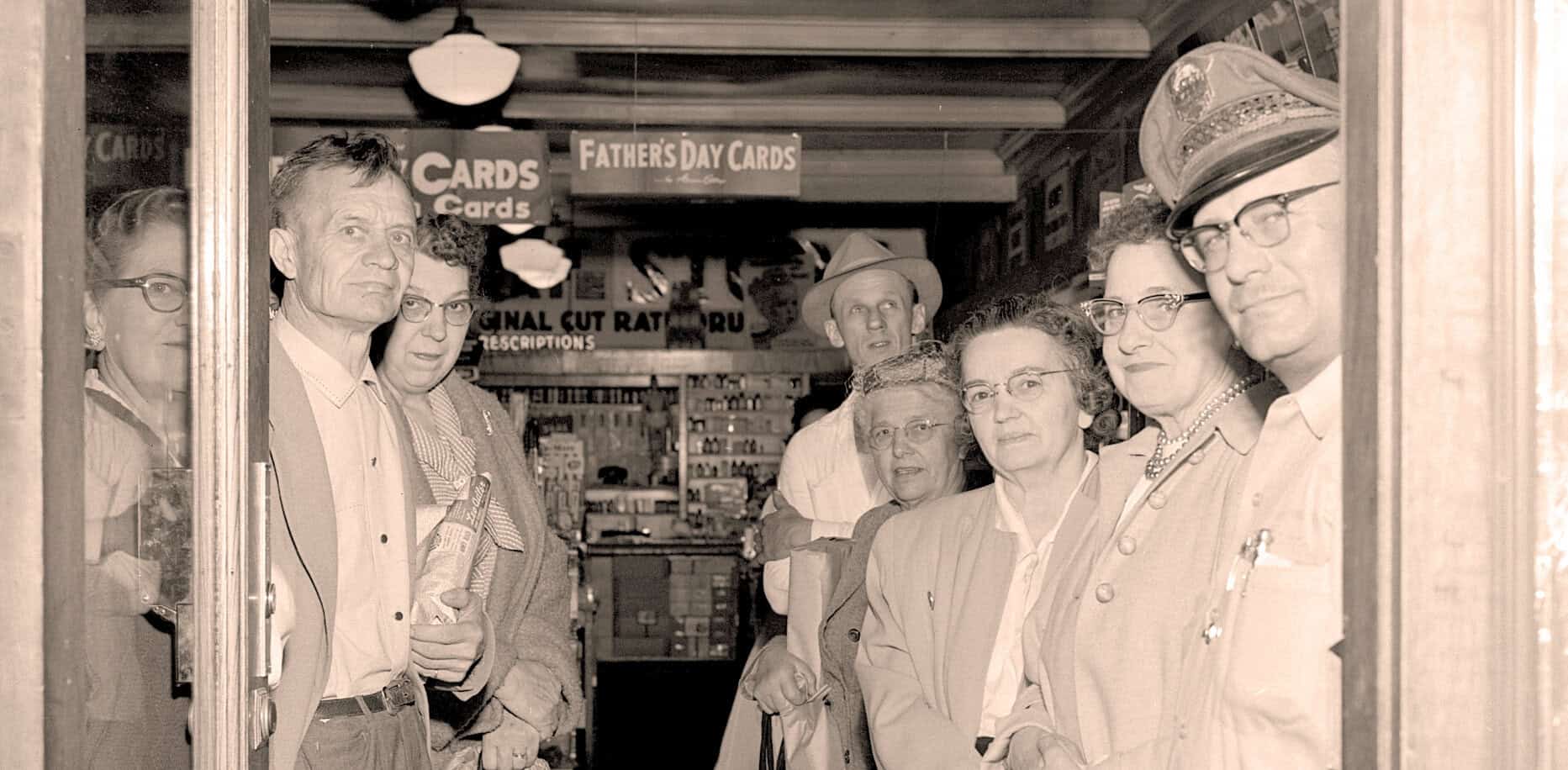 Vintage black and white photo of a group of people, including women and men wearing glasses and suits, inside a store with signs for Father's Day cards and other greeting cards, reflecting historical news and cultural moments.