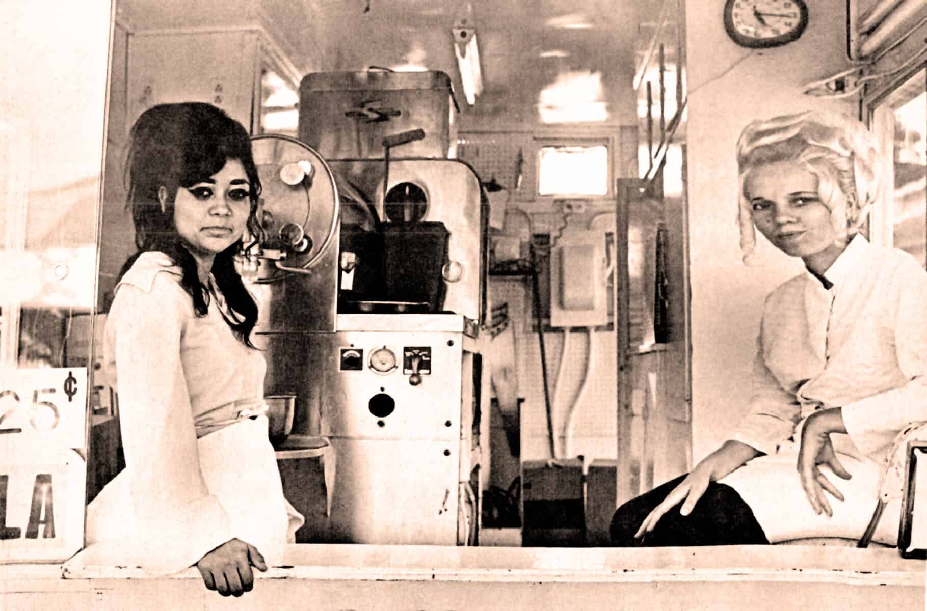 Vintage soda fountain workers, two women in classic uniforms, behind the counter at a nostalgic 1960s-style ice cream shop, showcasing historical food service and American culture.