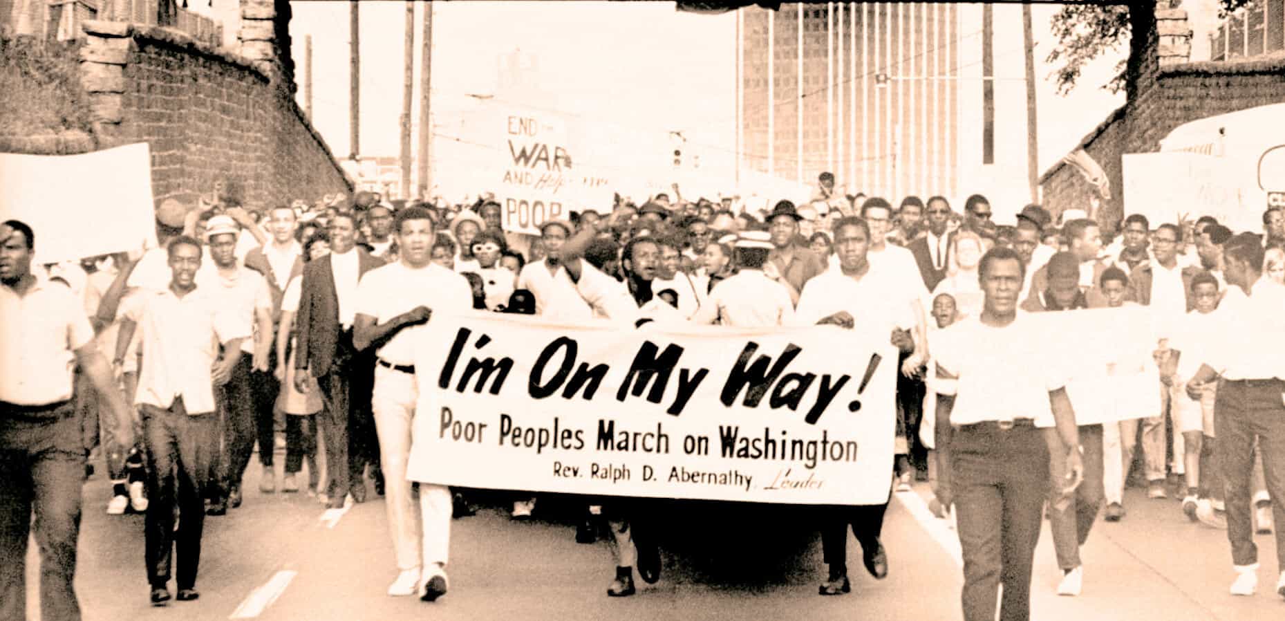 Protestors marching for civil rights during the Civil Rights Movement, holding a banner that reads "I'm On My Way! Poor Peoples March on Washington." In black and white, historic photo emphasizing news, history, and social activism.