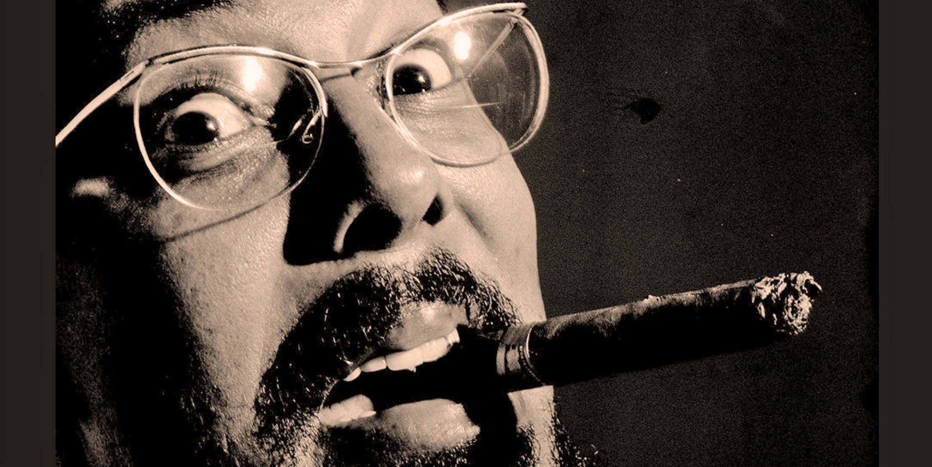 Close-up black and white photo of a man with glasses smoking a cigar, highlighting vintage style, news, history, and music archive themes.