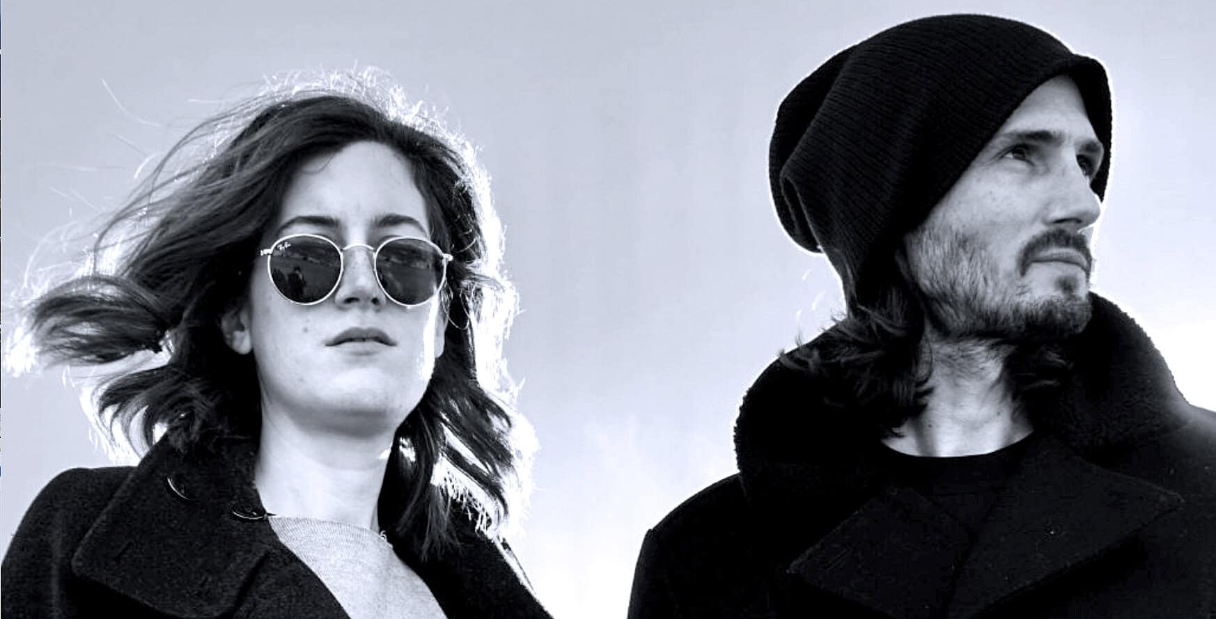 Vintage black and white photo of a young woman with wavy hair wearing sunglasses and a man with long hair and a beanie, both looking into the distance, representing music and cultural history for SEO.