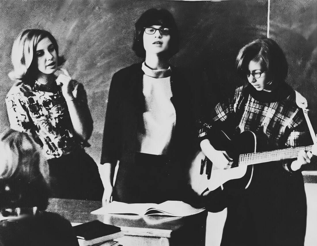 Black and white photo of three young women in a classroom scene, one playing guitar, capturing a nostalgic moment of youth, music, and education, suitable for historical and cultural archives.