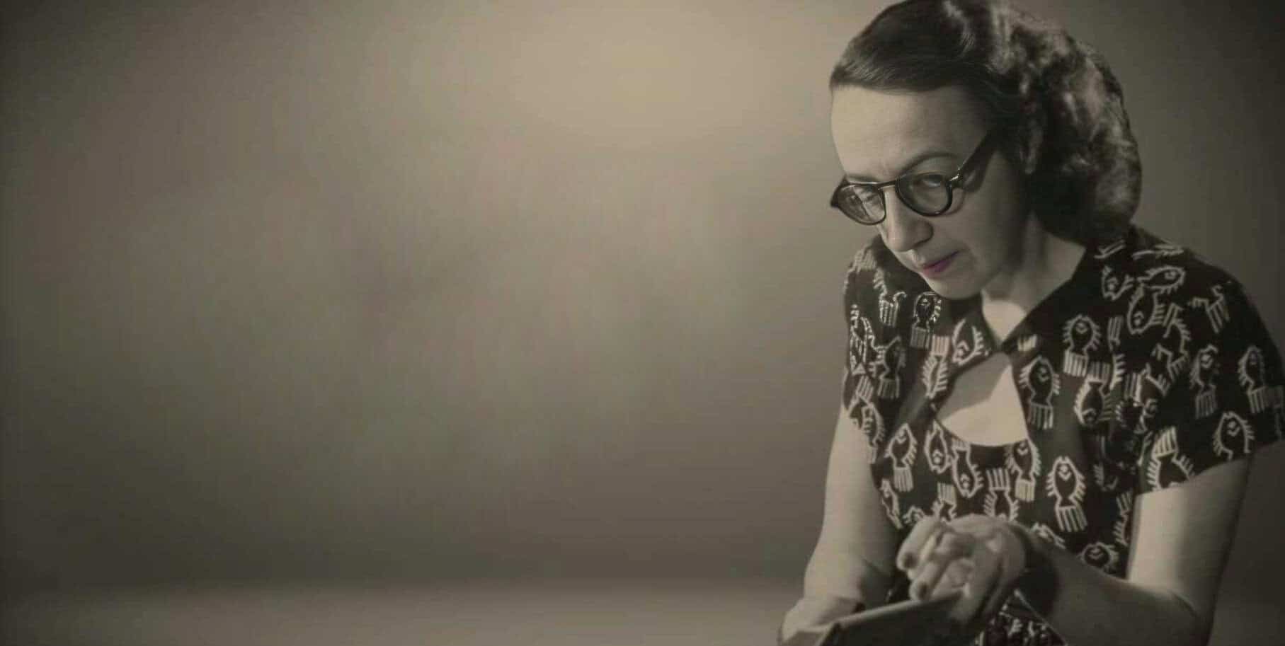 Vintage woman wearing glasses and a patterned dress, using a tablet against a plain background.