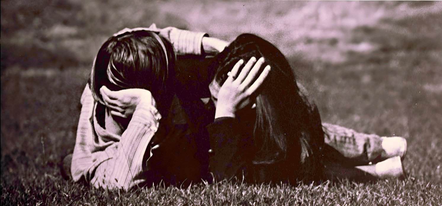Distressed young couple lying on grass during daytime, emotional support, comforting each other, heartbreak, emotional distress, outdoor setting, young adults, black and white, candid moment, personal struggle, mental health awareness.