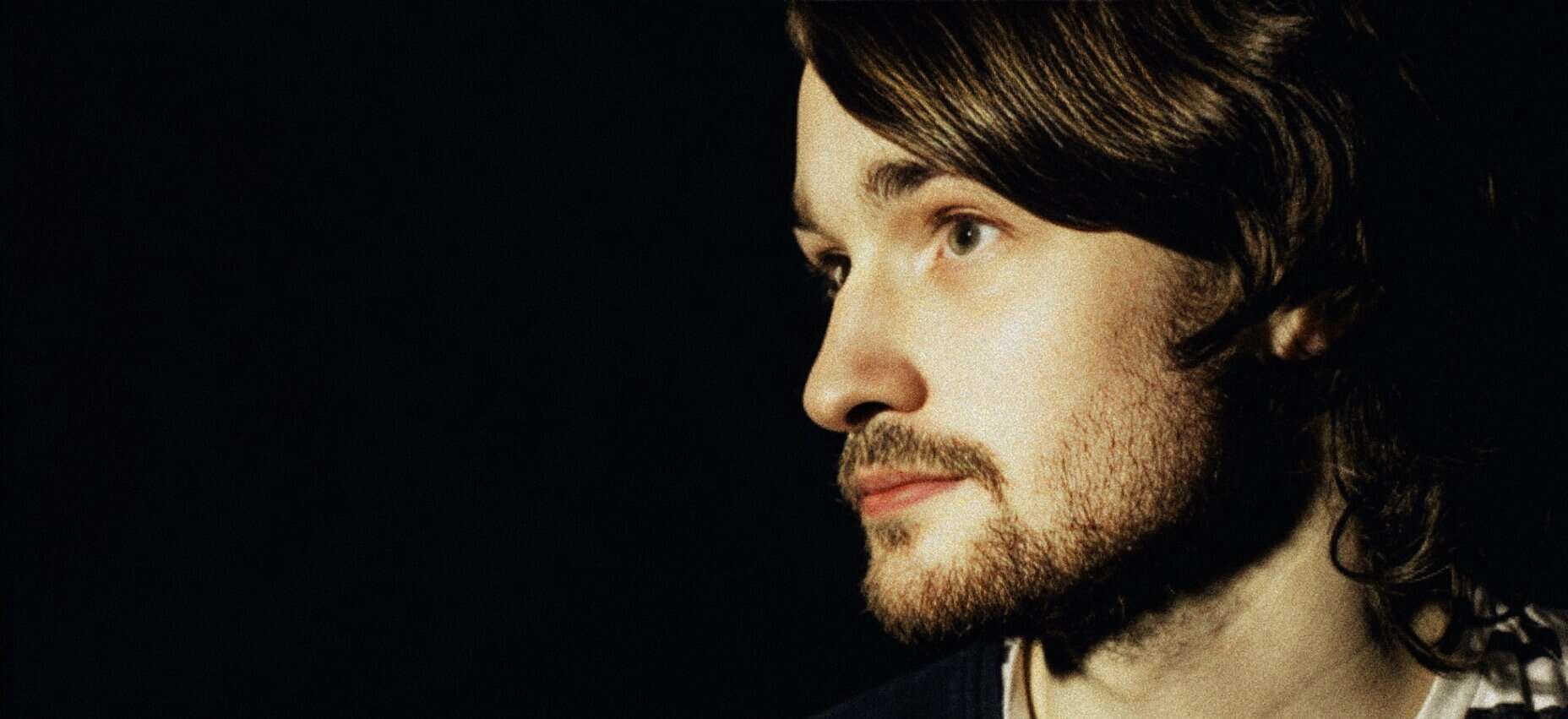 Close-up of a young man with long wavy hair and beard, thoughtful expression, dark background, emphasizing music, news, history, archival audio, and sound archive themes for SEO.