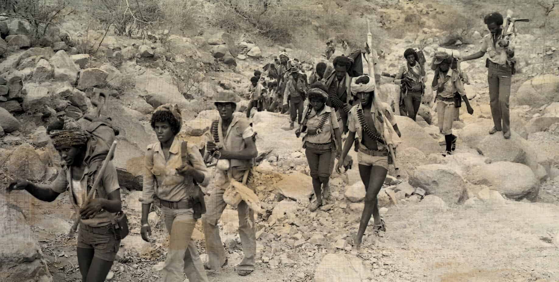 Children walking through a rocky, arid landscape carrying tools and supplies, depicting a scene of hardship and resilience in a rural or war-torn area, highlighting themes of struggle and survival.