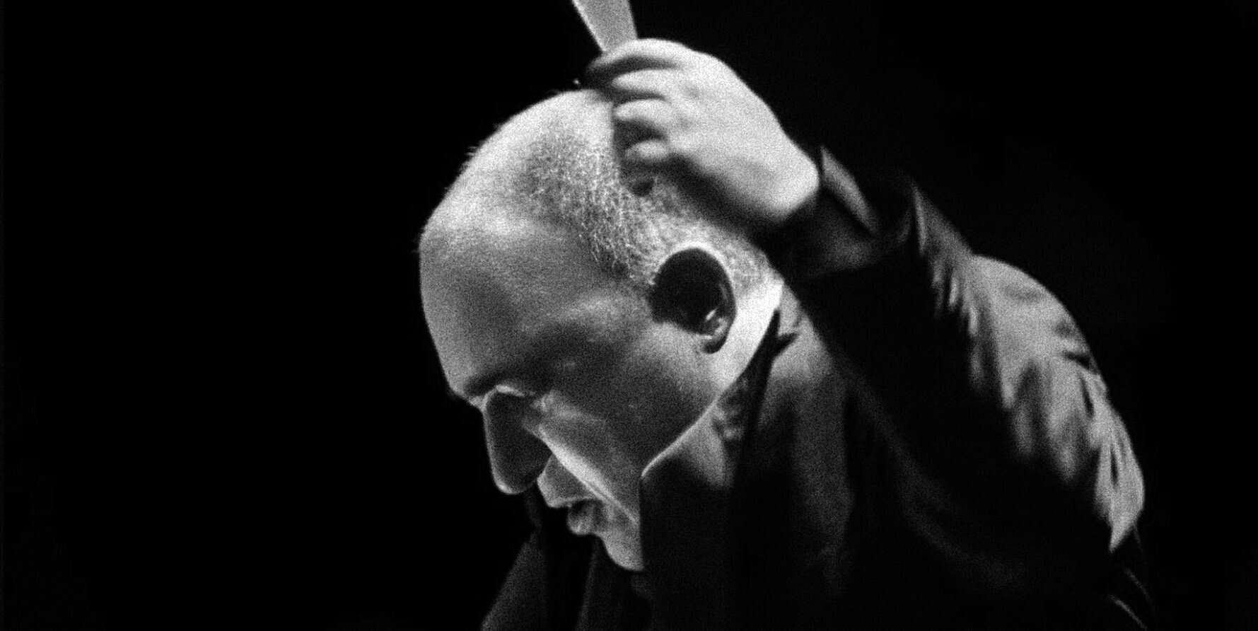 A black and white image of a man with a shaved head and beard, adjusting headphones, representing music, news, and sound archives on Past Daily.