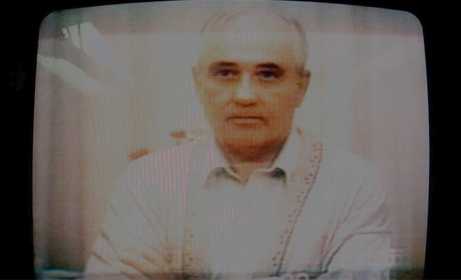 A vintage television screen displaying a black-and-white photo of a man with short hair, dressed in a white shirt, with a serious expression, referencing historic news, history, and music archives.