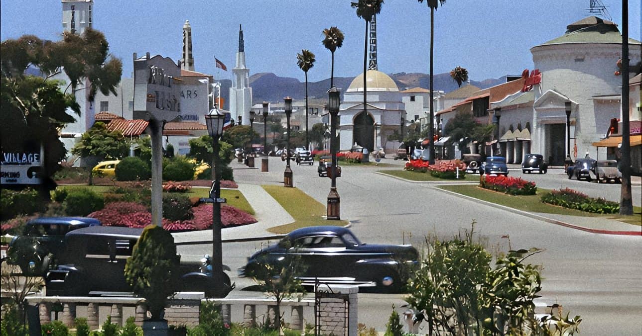 Vintage Hollywood-style cars driving through a sunny town square with palm trees and classic architecture, capturing mid-20th-century American city life.