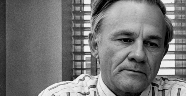 A black and white close-up of a man with light-colored hair, looking contemplative, with window blinds in the background, illustrating historical news, music, and media archives.