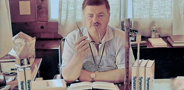 A man with a mustache holding glasses in a cluttered office, surrounded by books and papers, with a vintage workspace backdrop.