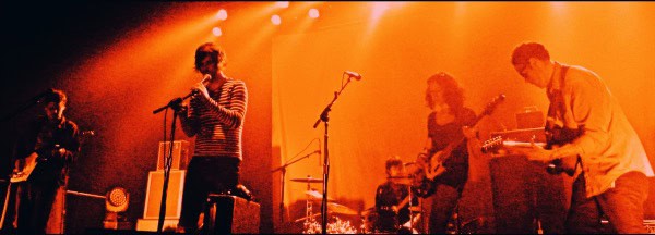 Live music performance with musicians playing guitars and singing on stage under warm orange lighting.