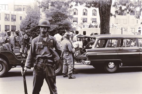 Police officer standing in front of a classic car during a historic event or protest, with other officers and spectators in the background. Black and white image capturing a moment of law enforcement history.