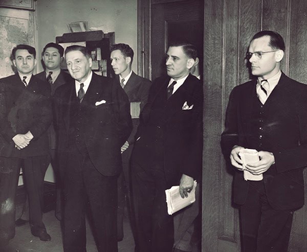 Male professionals in suits attending a formal meeting in an office setting, vintage black-and-white photo.