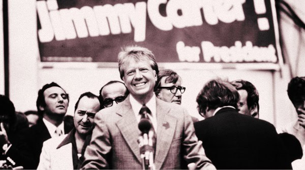 Bob Kennedy speaking at a political rally, capturing a historic moment in American politics with a backdrop featuring campaign signage and a diverse group of supporters.