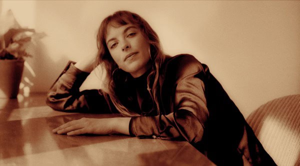 Vintage sepia-toned photograph of a young woman relaxing at a wooden table, reflecting nostalgic mood associated with historical news, music, and cultural archives from Past Daily.