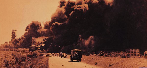 Devastating industrial fire with thick black smoke rising, vintage vehicles on the road, historical disaster scene emphasizing news, history, and sound archive of past events.