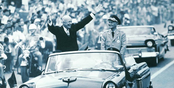 Prominent historical figures riding in a convertible during a public parade, capturing a significant moment in history with an enthusiastic crowd in the background.