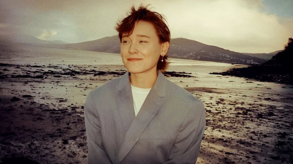 Vintage photograph of a young woman with short hair in a gray suit standing on a coastal beach with mountains in the background, capturing a nostalgic and peaceful moment.