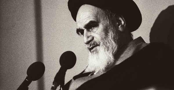 Historic black and white photo of a bearded man in traditional attire speaking at a microphone, symbolizing archival news and historical recordings from Past Daily.