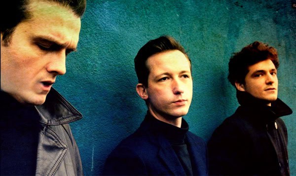 Desert Sessions band members standing against a vibrant blue wall, representing classic rock, alternative music, and music history.