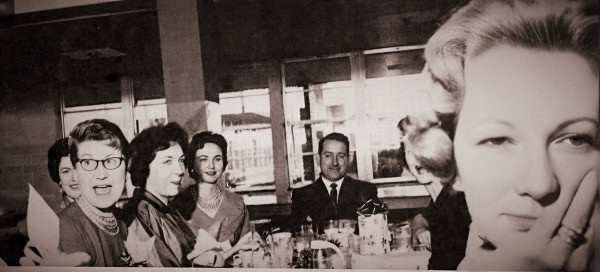 Vintage black and white photograph of a group of women and a man dining together in a mid-20th century restaurant or cafe, capturing social history and lifestyle.