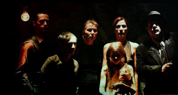 A group of five people, including children and adults, standing in a dimly lit room with a vintage atmosphere, representing themes of music, history, and nostalgia.