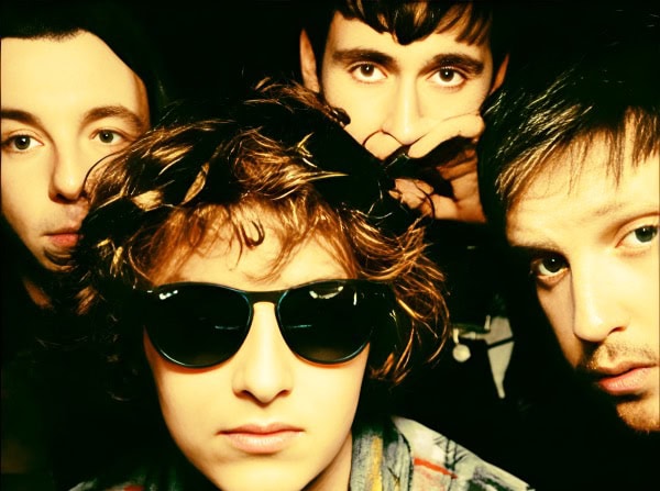 A band of four young adults with eclectic styles, including one wearing sunglasses, posing in a close-up shot for a music or news archive website.