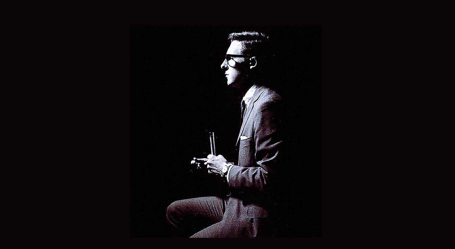 Vintage black and white image of a man in a suit and glasses holding a microphone, representing classic news, history, and music recordings from Past Daily sound archive.