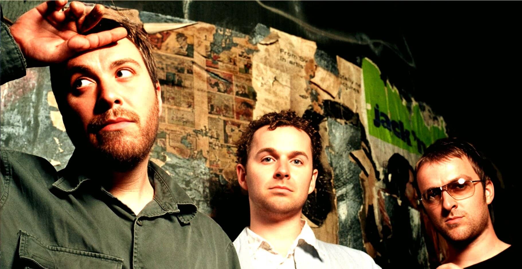 Vintage rock band photo featuring three male musicians posing in front of a collage wall with newspaper clippings and "numb" graffiti, emphasizing music history and alternative band culture, perfect for music archive content.