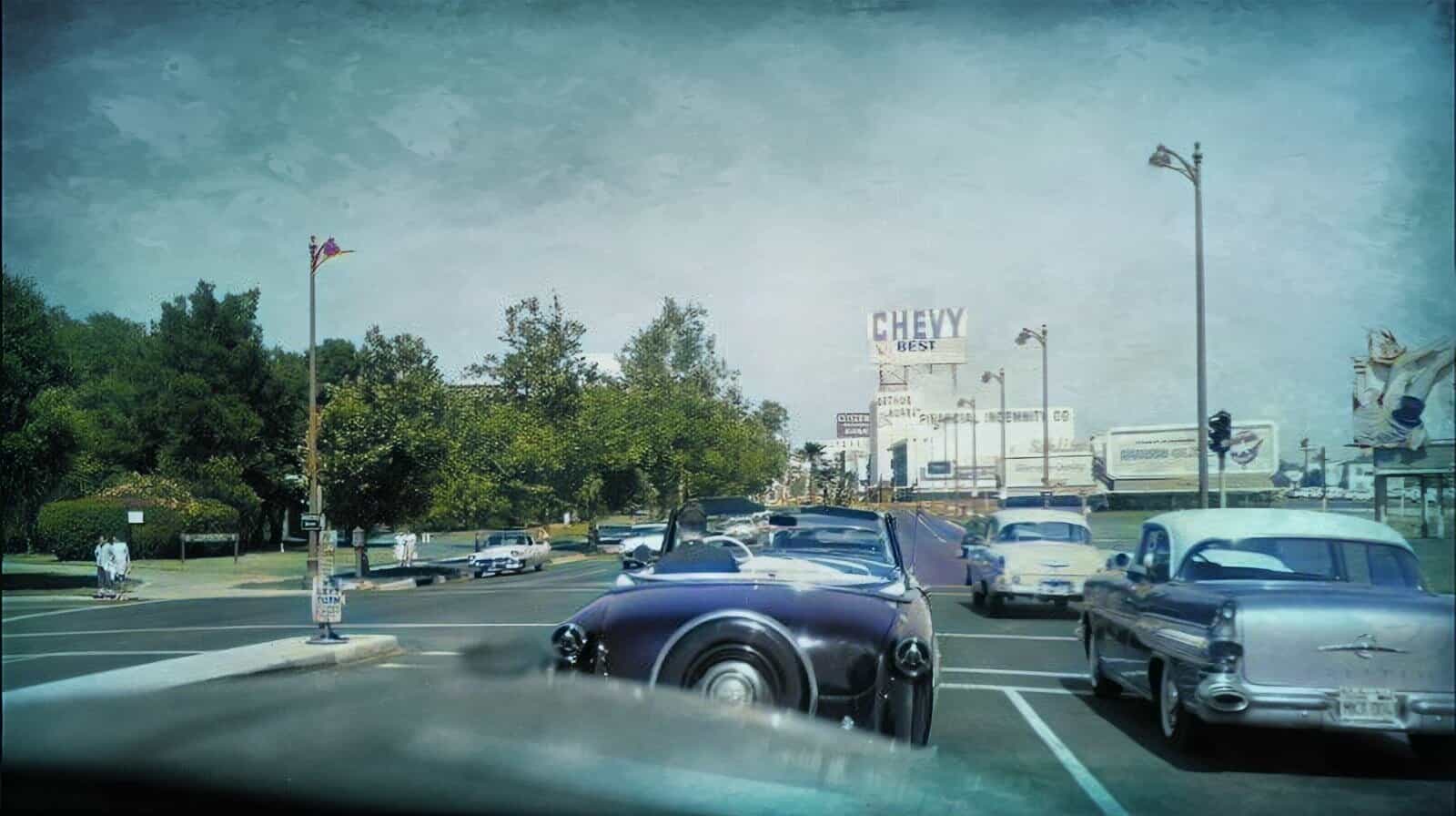 Vintage convertible car parked on a busy city street with historic buildings and billboards in the background, capturing mid-20th century American urban scene.
