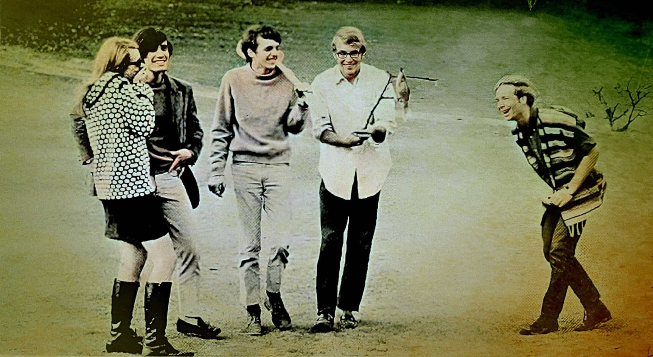 Vintage group of five young people on a film set or photo shoot, dressed in 1960s casual fashion, engaging in lively conversation; nostalgic scene capturing youth culture and creative expression.