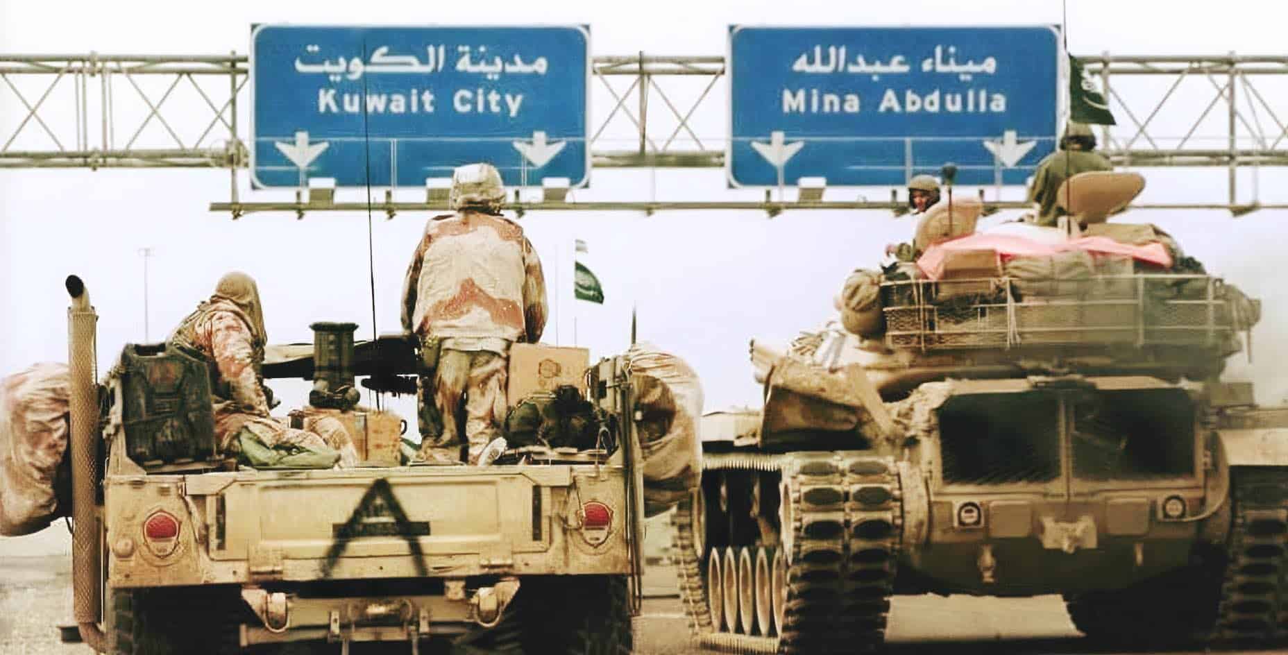 Military tanks with soldiers on it on a highway in Iraq, with signs indicating Kuwait City and Mina Abdulla, during a conflict or military operation.