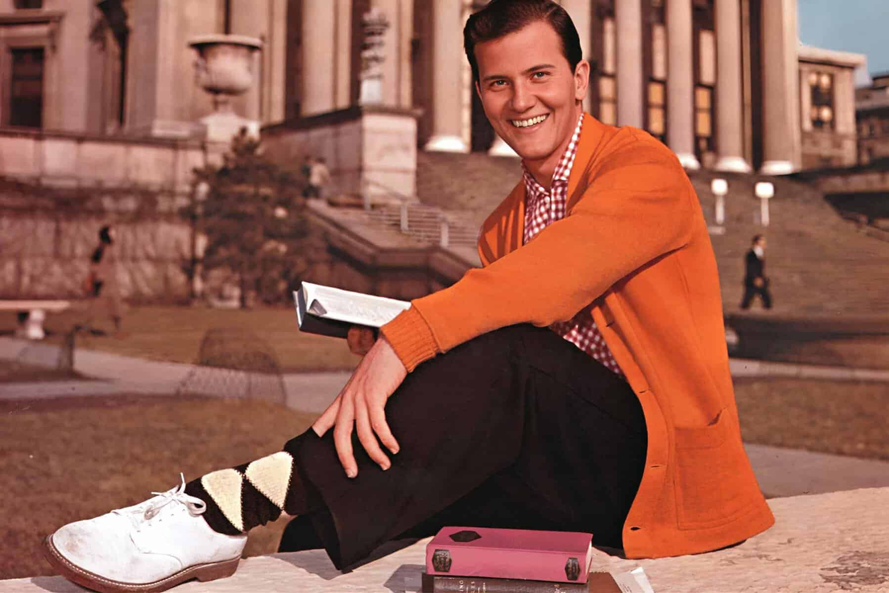 An orange-jacketed man sitting on steps outside a historic building, holding a book, smiling, with a pink book lying nearby, capturing a classic scene reminiscent of "The Breakfast Club" movie.