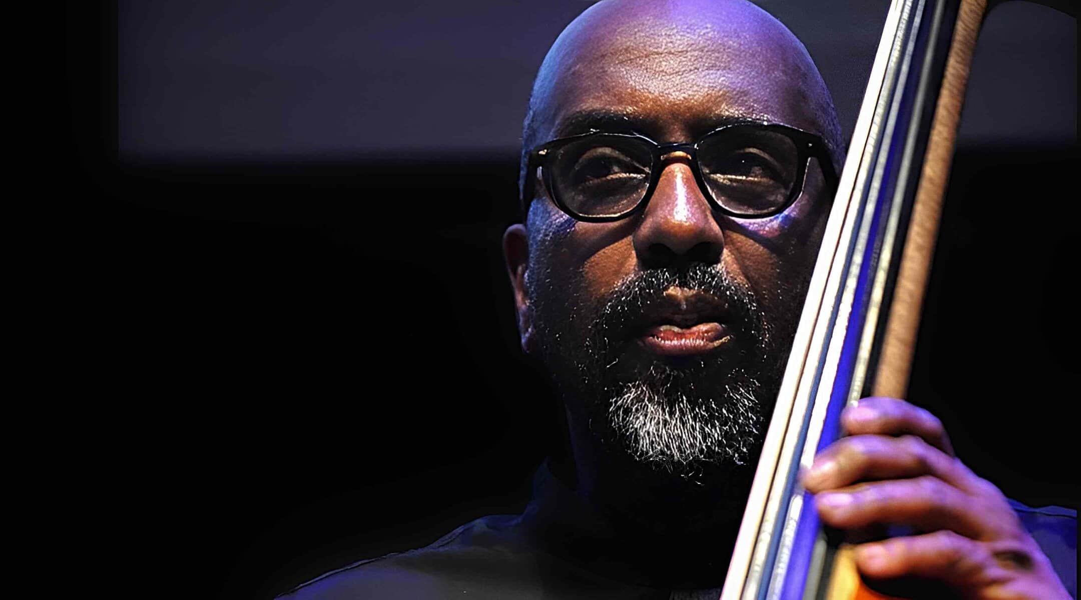 Bassist playing jazz music with a serious expression, captured in a close-up shot highlighting his face, glasses, and the neck of his bass guitar.