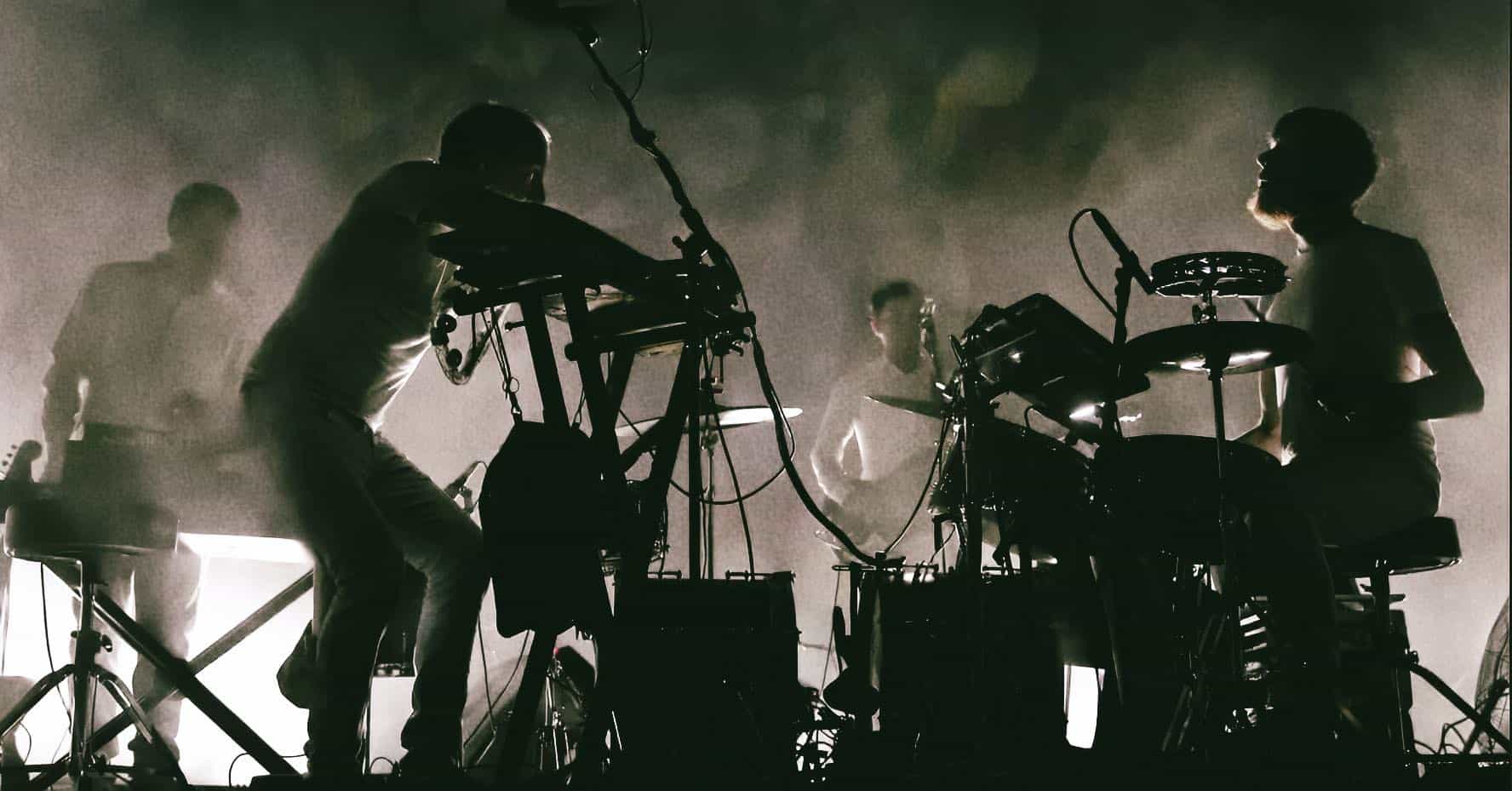 Dynamic electronic music performance featuring musicians with electronic instruments on stage, silhouetted against smoky lighting effects.