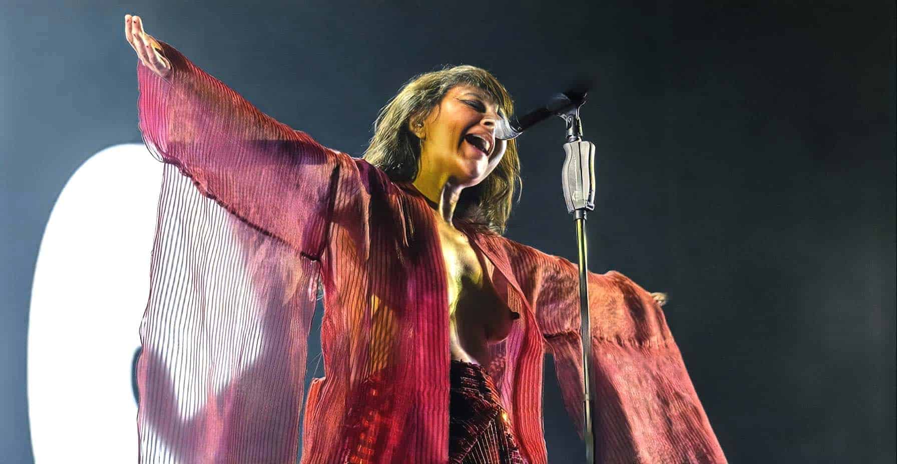 Vintage female singer passionately performing on stage with arms outstretched, wearing a flowing red and pink dress, under dramatic lighting for a music and news archive.
